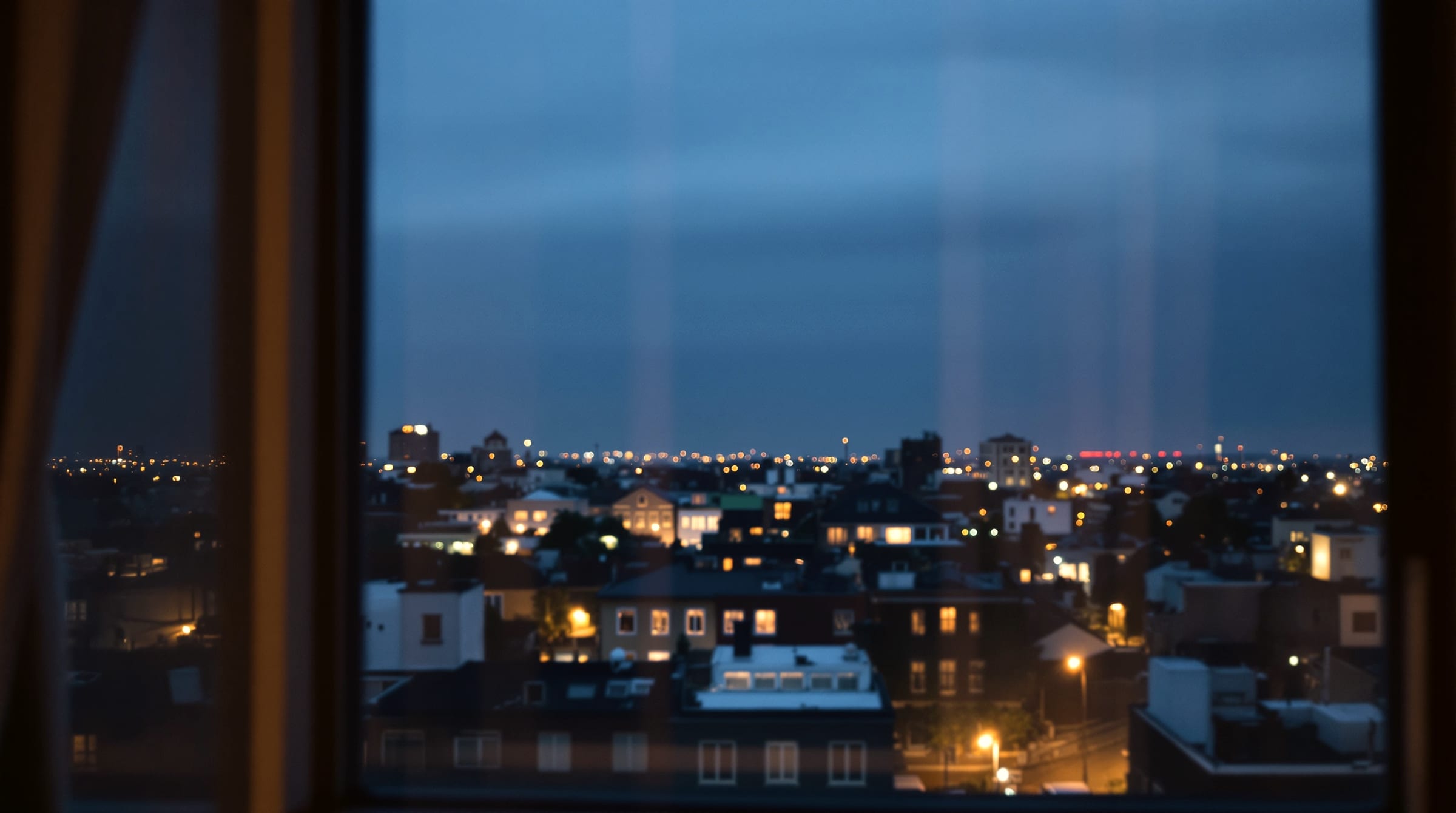 An urban view through a window at blue hour, anonymous rooftops and scattered warm lights, no recognizable skyline